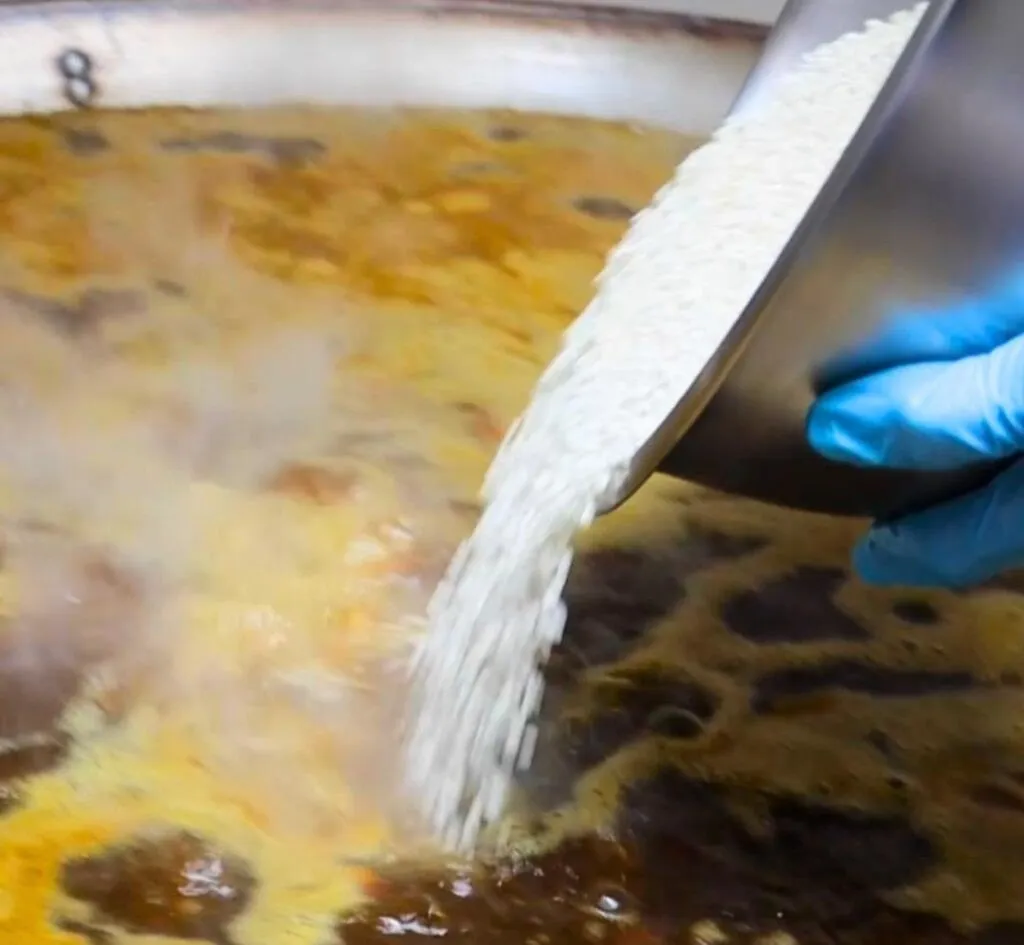 Arroz cayendo sobre el caldo de una paellera grande en el obrador de Xepep