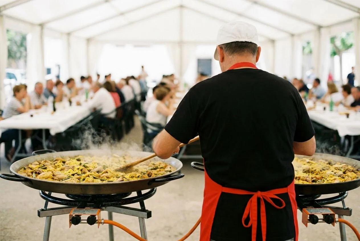Dos paellas grandes terminándose bajo una carpa, con un cocinero de espaldas y los invitados sentados al fondo